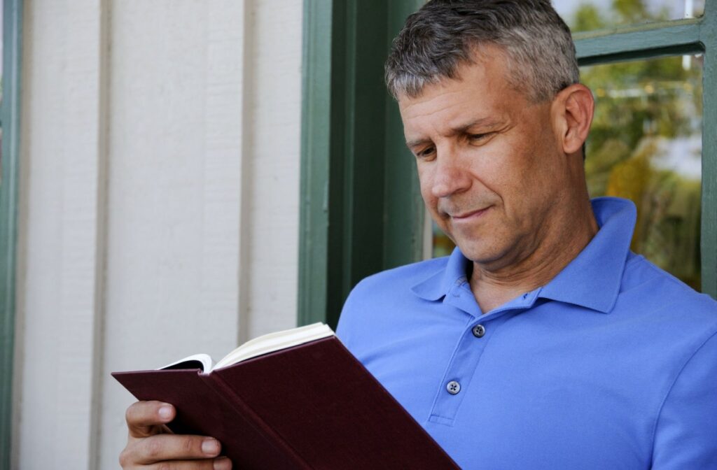 A middle-aged man not wearing glasses sitting outside by a window and reading a book.