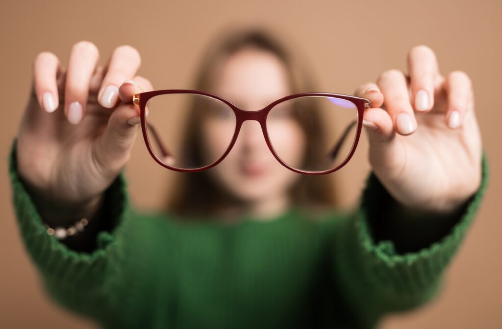 A blurry, backgrounded figure holds out a pair of glasses with maroon frames.