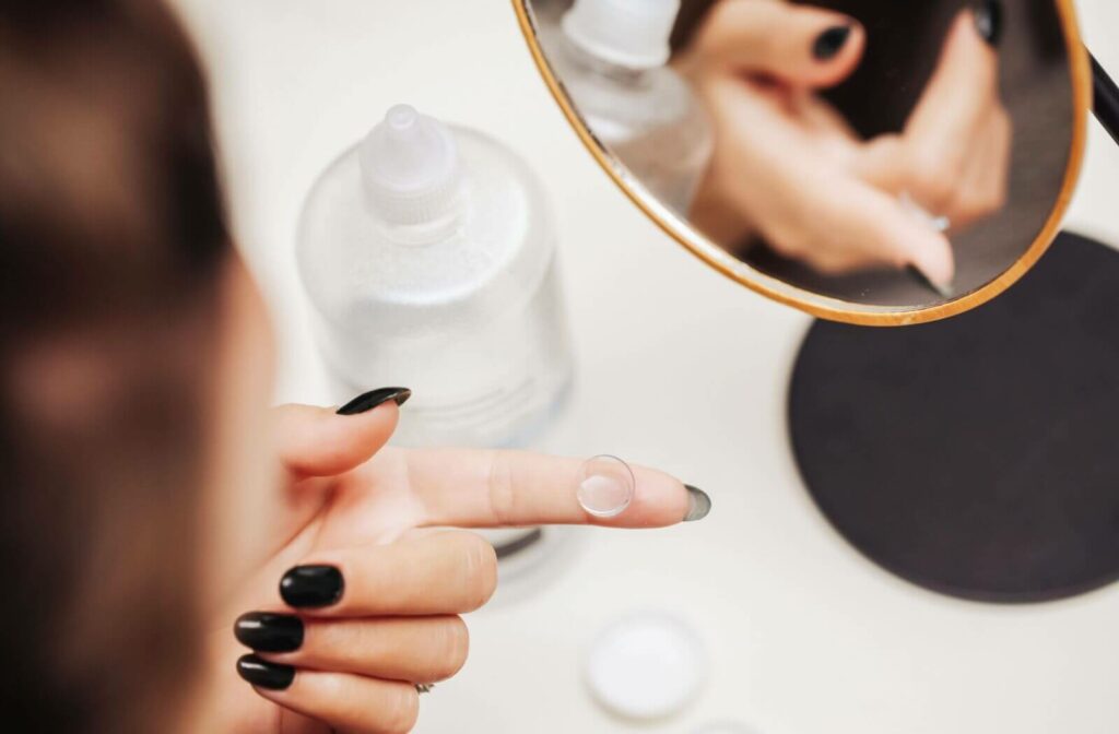 A person holding a contact lens on their left index finger as it's reflected in a small cosmetic mirror on a bathroom counter