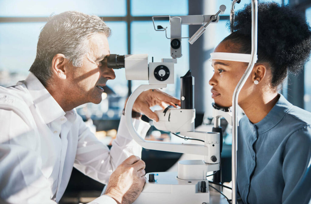 An optometrist smiling while carefully examining a young patient's eyes during an eye exam in a sunlit optometry office.