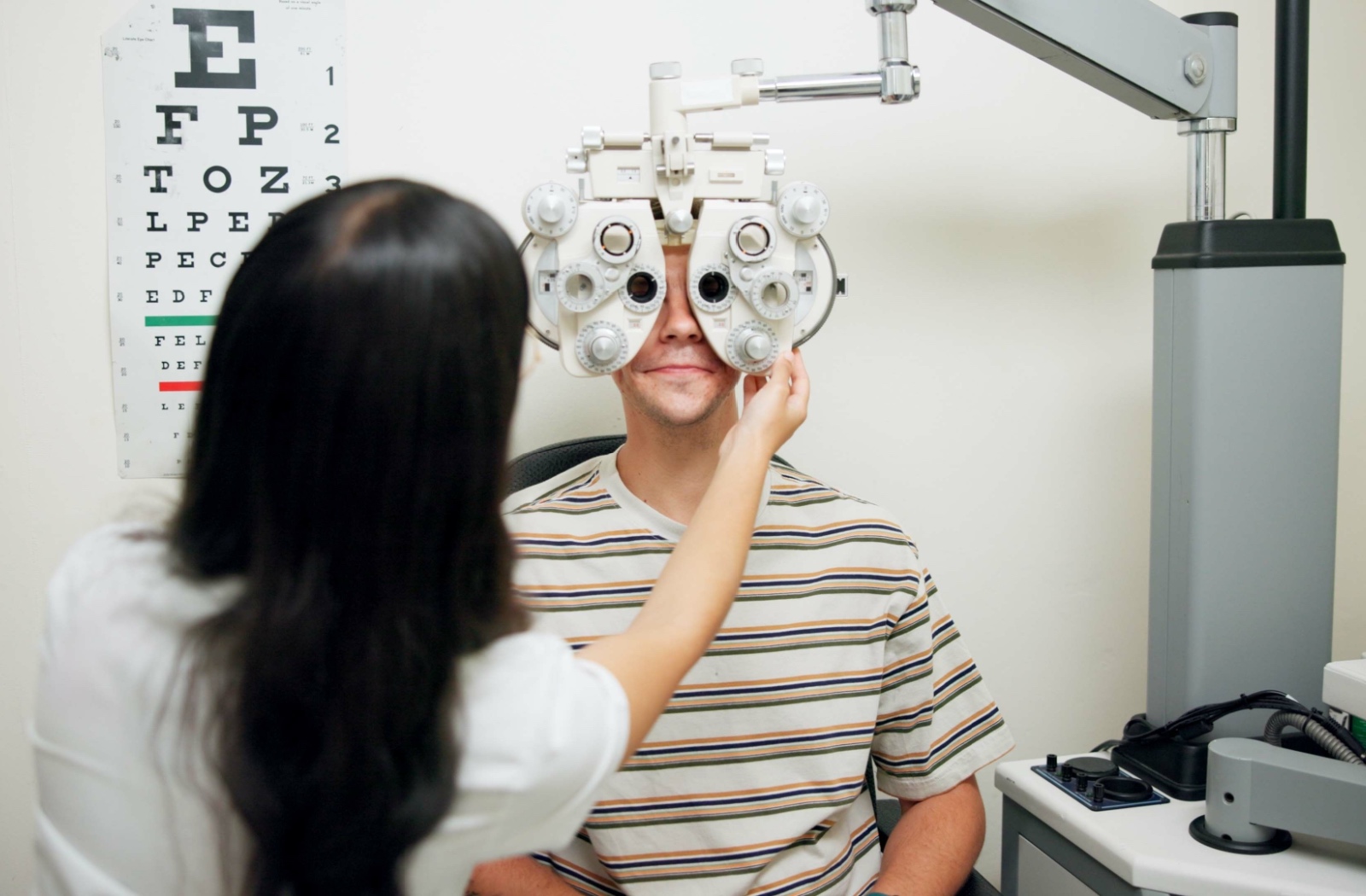 Adult undergoing an eye exam with a large diagnostic machine in front of their face as the optometrist adjusts the equipment.