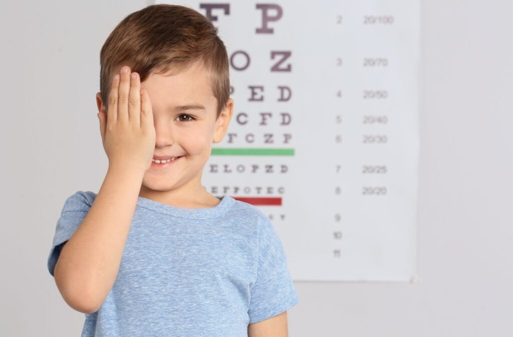 A child smiles and covers one eye while standing in front of an eye exam.