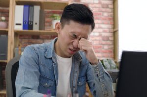 Young adult sitting at a desk rubbing their left eye with irritated or painful expression.